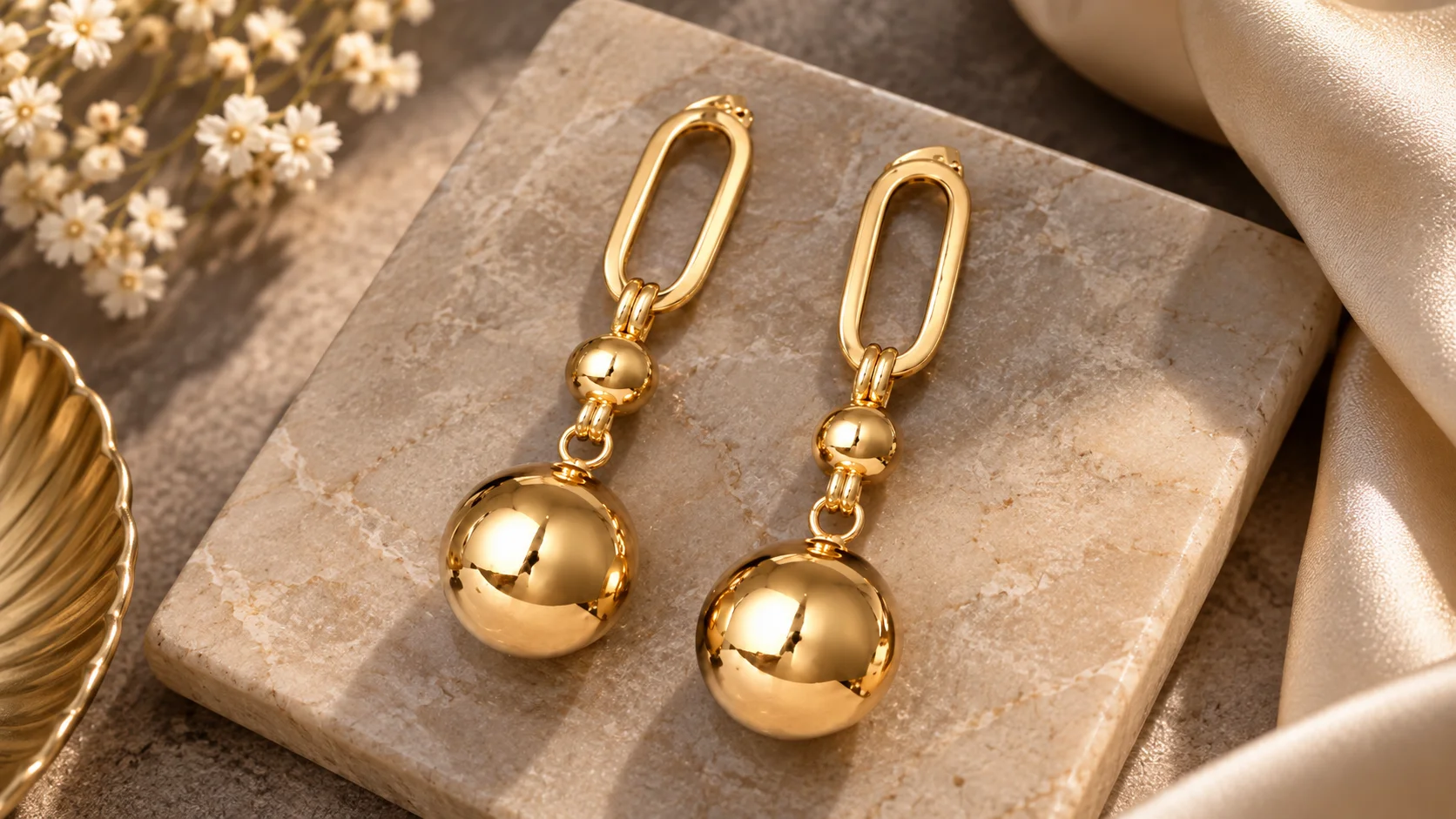 Gold-tone drop earrings with elongated geometric loops and polished spherical drops styled in an elegant jewellery editorial setting.