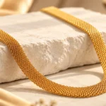 A gold-tone plated mesh necklace arranged in a curved shape on a cream stone display, with a warm ivory and champagne-beige background. Gold mesh necklace styling inspiration for neutral outfits, everyday jewellery and occasion wear.