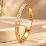 Gold-tone plated bangle with clear round decorative stones and textured gold borders displayed on champagne-beige props.