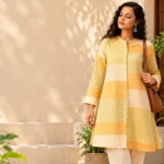 Yellow striped tunic styling with mustard-yellow and cream colour-blocked panels, pale sage stripes and a flared silhouette, styled against a warm neutral background with green plants.